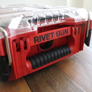 May include: A red and black tool case with a clear lid. The text "RIVET GUN" is printed in black on a red handle. The case has a black, ribbed handle and metal hinges.