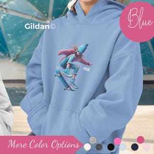 May include: A light blue hoodie featuring a graphic of a person skateboarding. The graphic includes the name "MIA". The word "Blue" is written in cursive on a pink circle. The hoodie has a front pocket and a hood. The brand name "Gildan" is visible.