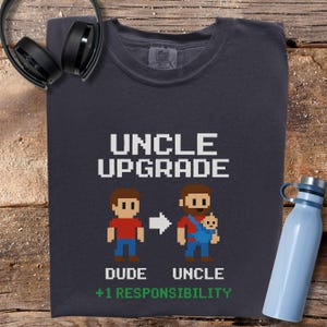Pode incluir: Camiseta cinza escuro com o texto "UNCLE UPGRADE" em letras pixeladas brancas. Abaixo, duas figuras pixeladas, uma com a etiqueta "DUDE" e a outra "UNCLE" segurando um bebê. O texto "+1 RESPONSIBILITY" está abaixo.