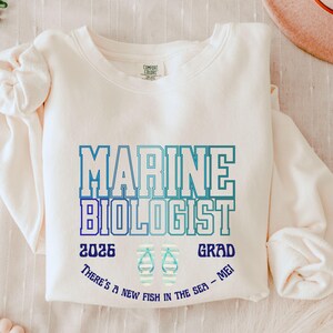 May include: A cream-coloured sweatshirt featuring the text "MARINE BIOLOGIST" in blue and teal block letters. The numbers "2026" and "GRAD" are printed below, along with a flip-flop graphic. The phrase "THERE'S A NEW FISH IN THE SEA - ME!" is printed below.