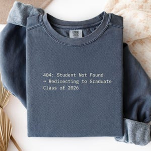 Op de afbeelding: Een leisteenblauwe Comfort Colors sweatshirt met de tekst "404: Student Not Found -> Redirecting to Graduate Class of 2026" in het wit. De sweatshirt heeft een ronde hals en lange mouwen.