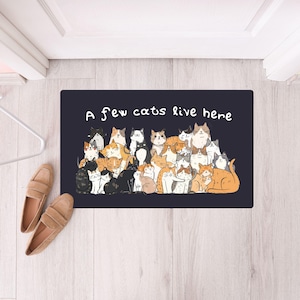Könnte beinhalten: Eine rechteckige Fußmatte in Marineblau mit einer Cartoon-Illustration zahlreicher Katzen in verschiedenen Posen und Farben. Der weiße Text "A few cats live here" steht oben. Beige Loafer sind links.