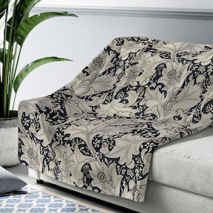 Pode incluir: Uma manta creme com um padrão floral azul marinho escuro drapeada sobre um sofá branco. O design botânico apresenta folhas grandes e elementos florais menores. A manta adiciona um toque decorativo à sala de estar.