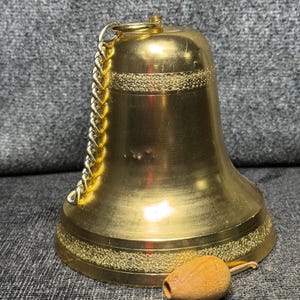 May include: A polished gold-tone brass bell featuring a textured horizontal band near the top and bottom. A gold-colored metal chain is attached to the top loop, and a small, light brown wooden clapper hangs from the bottom by a thin cord.