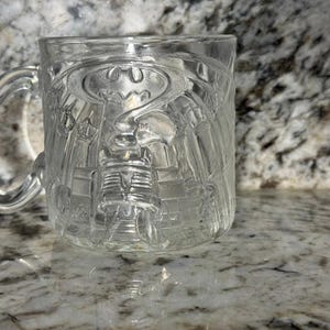 May include: A clear glass mug featuring a raised, embossed design of the Batman logo and a cityscape. The mug has a curved handle on the left side and a smooth, transparent finish, showcasing the detailed molded texture of the superhero emblem.