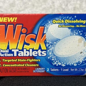 Puede incluir: Una caja rectangular de tabletas de detergente para ropa Wisk Dual Action. El empaque es rojo y azul con texto amarillo que dice NEW!, Quick Dissolving, No Measuring... No Mess y Targeted Stain-Fighters. Peso neto 76,5g.