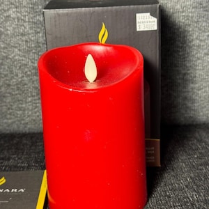 May include: A bright red pillar candle with a realistic LED flickering flame. The candle has a slightly curved top and is in front of a black box with a yellow flame graphic. A small card is visible.