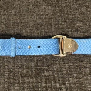 May include: A light blue belt with a textured snakeskin pattern. The belt features a silver-colored buckle with a decorative emblem. The belt is made of a durable material and has several holes for adjustment.