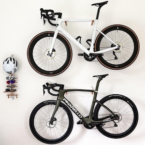 Peut inclure: Deux vélos de route montés sur un mur blanc. L'un est blanc avec des accents noirs, l'autre est vert olive avec des composants noirs. Les deux ont des roues noires avec des flancs beiges. Un casque et des lunettes de soleil sont également montés sur le mur.