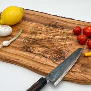 Op de afbeelding: Een houten snijplank met een ingebrand ontwerp van vlammen en een grill. De plank heeft een natuurlijke houtkleur en is ovaalvormig. Er liggen verschillende groenten op de plank, waaronder een citroen, knoflook, een groene ui, tomaten en een gele paprika. Een mes met een zwarte handgreep ligt ook op de plank.