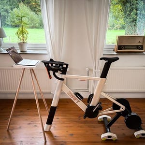 Könnte beinhalten: Ein weiß-schwarzes Indoor-Trainingsfahrrad mit orangefarbenen Akzenten und dem Schriftzug "ZWIFT". Ein Laptop steht auf einem kleinen Holztisch. Ein Vintage-Radio steht auf einer Fensterbank.