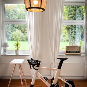 Op de afbeelding: Indoor hometrainer met het woord "ZWIFT" op het frame. De fiets is wit en zwart, met een zwart zadel en stuur. De fiets staat op een houten vloer, met een witte muur en een raam op de achtergrond.