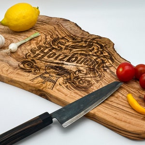 May include: A wooden cutting board with a natural wood grain pattern. The board has a burnt-in design that says "Holzbude" with a grill image. The board is surrounded by a lemon, garlic, green onions, tomatoes, and a yellow pepper. A black handled knife is also on the board.