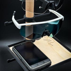 May include: A display stand with two pairs of sports sunglasses and a smartphone. The sunglasses have coloured lenses and white or green frames. The phone is black and grey. The stand is black with a wooden base.