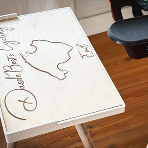 May include: A light-colored wooden table with a white frame and a map design. The table has the words "Dasde Beste Cycling" and the name "Emil" engraved on it. The table is next to a stationary exercise bike.