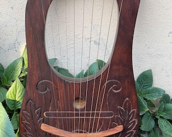 May include: A wooden lyre with a brown stain and intricate carvings. The lyre has 10 strings and a wooden bridge. The lyre is sitting on a concrete surface.