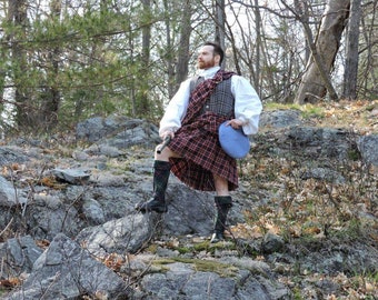 Peut inclure: Un homme portant un kilt à carreaux, une chemise blanche et un gilet se tient sur une colline rocheuse. Il tient un béret bleu dans sa main droite et un bâton dans sa main gauche. Il regarde le ciel.