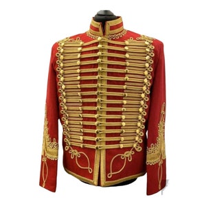 Puede incluir: Una chaqueta roja con ribetes y botones dorados. La chaqueta tiene un cuello alto y mangas largas. Los ribetes están en un patrón trenzado y los botones son redondos y dorados.