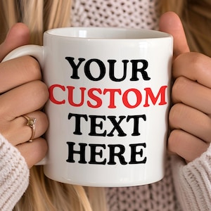 May include: White ceramic mug with the text "YOUR CUSTOM TEXT HERE" in black and red. The mug is held by a person wearing a cream-coloured sweater. The mug has a handle on the right side.