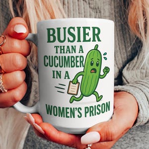 Puede incluir: Una taza de cerámica blanca con un personaje de pepino de dibujos animados verde corriendo con un portapapeles. El texto dice: BUSIER THAN A CUCUMBER IN A WOMEN'S PRISON. El diseño está impreso en letras verde oscuro en la parte frontal.