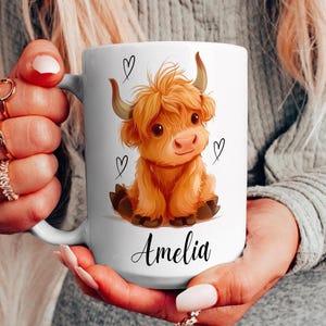 Pode incluir: Caneca de cer&acirc;mica branca com uma ilustra&ccedil;&atilde;o de desenho animado de uma vaca Highland. A vaca tem um casaco laranja fofo, chifres pequenos e detalhes de cora&ccedil;&atilde;o. O nome "Amelia" est&aacute; escrito abaixo.