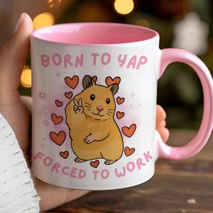 May include: White ceramic mug with a pink interior and handle. The mug features a cartoon hamster giving a peace sign, surrounded by hearts, and the text "BORN TO YAP FORCED TO WORK".
