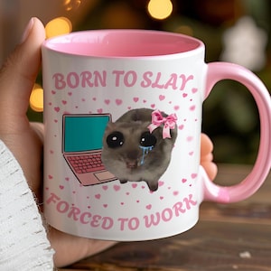 May include: White ceramic mug with a pink interior and handle. The mug features the text "BORN TO SLAY FORCED TO WORK" with a cartoon illustration of a crying animal wearing a pink bow and a laptop. Pink hearts are scattered around the design.