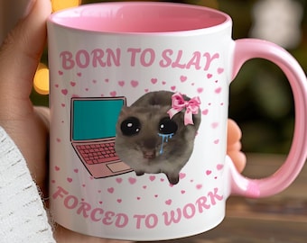 May include: White ceramic mug with a pink interior and handle. The mug features the text "BORN TO SLAY FORCED TO WORK" with a cartoon illustration of a crying animal wearing a pink bow and a laptop. Pink hearts are scattered around the design.