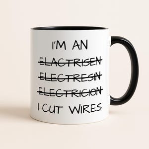 Op de afbeelding: Witte keramische mok met een zwart handvat en rand. De mok heeft de tekst "I'M AN ELECTRICIAN I CUT WIRES" in het zwart, met de woorden "ELACTRISEN", "ELECTRESIN" en "ELECTRICION" doorgestreept.