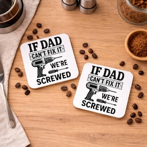 May include: Two square white coasters with the text "IF DAD CAN'T FIX IT WE'RE SCREWED" in black. Each coaster features a black illustration of a drill and a screwdriver. Coffee beans and other items are scattered around the coasters.