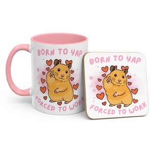 May include: A white ceramic mug with a pink interior and handle, and a matching square coaster. Both feature a cartoon hamster with a peace sign, surrounded by hearts, and the text "BORN TO YAP FORCED TO WORK."