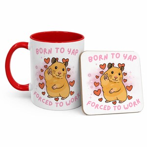 May include: A white ceramic mug with a red interior and handle, and a matching coaster. Both feature a cartoon hamster giving a peace sign, surrounded by hearts, with the text "BORN TO YAP FORCED TO WORK."