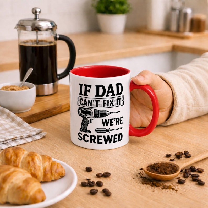 May include: White coffee mug with a red interior and handle, featuring the text "IF DAD CAN'T FIX IT WE'RE SCREWED" along with a drill and screwdriver graphic. A French press, croissants, and coffee beans are also visible.