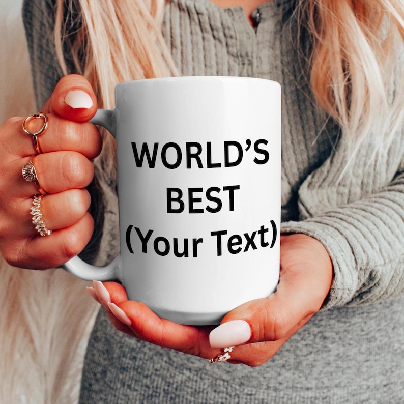 Puede incluir: Una taza de cer&aacute;mica blanca con el texto "WORLD'S BEST (Your Text)" en negro. La taza est&aacute; sostenida por una persona que lleva un jersey gris. La taza tiene un asa en el lateral.