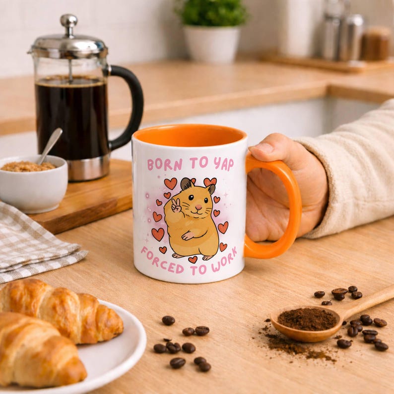 May include: White ceramic mug with an orange interior and handle, featuring a cartoon hamster with a peace sign and the text "BORN TO YAP FORCED TO WORK". The mug is surrounded by heart graphics.