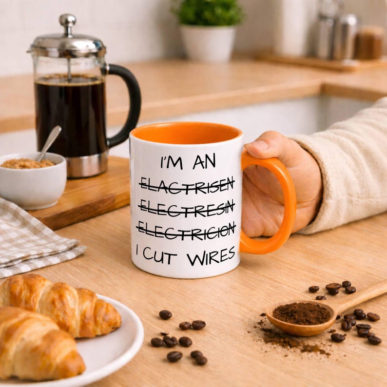 May include: A white ceramic mug with an orange interior and handle, featuring black text that reads: I'M AN, ELACTRISEN, ELECTRESIN, ELECTRICION, I CUT WIRES. The words ELACTRISEN, ELECTRESIN, and ELECTRICION are crossed out with a horizontal black line.
