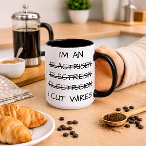 May include: A white ceramic mug with a black interior and handle, featuring the black text: I'M AN, ELACTRISEN, ELECTRESIN, ELECTRICION, I CUT WIRES. The words ELACTRISEN, ELECTRESIN, and ELECTRICION are crossed out with a horizontal black line.