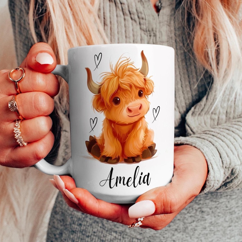 Pode incluir: Caneca de cer&acirc;mica branca com uma ilustra&ccedil;&atilde;o de desenho animado de uma vaca Highland. A vaca tem um casaco laranja fofo, chifres pequenos e detalhes de cora&ccedil;&atilde;o. O nome "Amelia" est&aacute; escrito abaixo.
