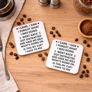 K&ouml;nnte beinhalten: Zwei quadratische wei&szlig;e Untersetzer mit humorvollem Text, darunter "I CAME, I SAW, I FORGOT WHAT I WAS DOING". Kaffeebohnen und kleine Schalen sind auf einer Holzoberfl&auml;che um die Untersetzer herum verstreut.