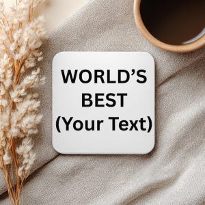 Puede incluir: Un posavasos cuadrado blanco con esquinas redondeadas, con el texto "WORLD'S BEST (Your Text)" en negro. El posavasos est&aacute; sobre una superficie de tela gris, con una taza de caf&eacute; y flores secas en el fondo.