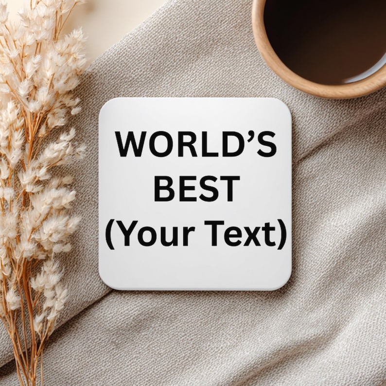 Puede incluir: Un posavasos cuadrado blanco con esquinas redondeadas, con el texto "WORLD'S BEST (Your Text)" en negro. El posavasos est&aacute; sobre una superficie de tela gris, con una taza de caf&eacute; y flores secas en el fondo.
