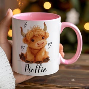 Pode incluir: Caneca de cer&acirc;mica branca com interior e al&ccedil;a rosa. A caneca apresenta uma ilustra&ccedil;&atilde;o de desenho animado de uma vaca Highland com cora&ccedil;&otilde;es e o nome "Mollie" em escrita preta. A vaca &eacute; marrom com pequenos chifres.