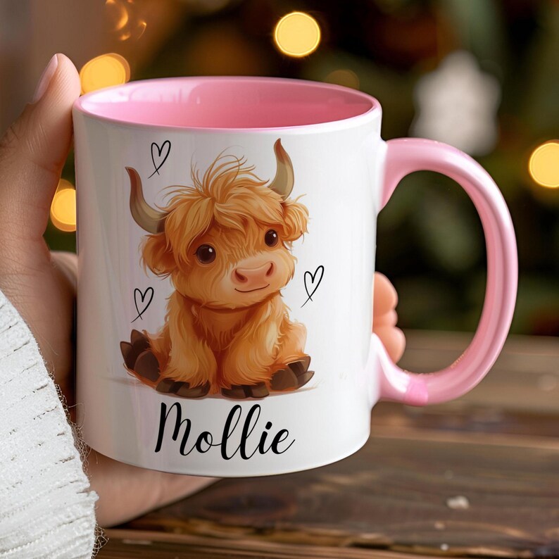 Pode incluir: Caneca de cer&acirc;mica branca com interior e al&ccedil;a rosa. A caneca apresenta uma ilustra&ccedil;&atilde;o de desenho animado de uma vaca Highland com cora&ccedil;&otilde;es e o nome "Mollie" em escrita preta. A vaca &eacute; marrom com pequenos chifres.