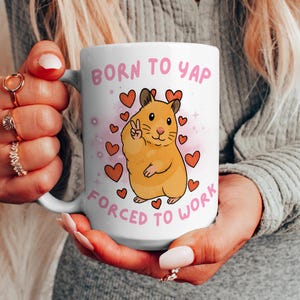 May include: White ceramic mug with the text "BORN TO YAP FORCED TO WORK" in pink. The mug features a cartoon hamster giving a peace sign, surrounded by pink hearts.