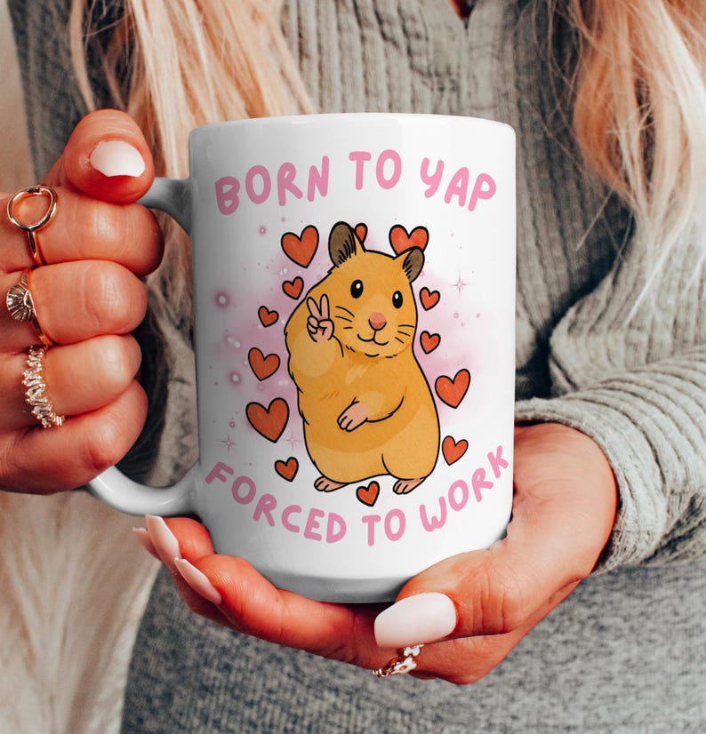 May include: White ceramic mug with the text "BORN TO YAP FORCED TO WORK" in pink. The mug features a cartoon hamster giving a peace sign, surrounded by pink hearts.