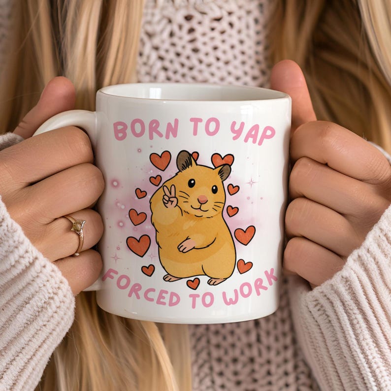 May include: White ceramic mug with the text "BORN TO YAP FORCED TO WORK" in pink. The mug features a cartoon hamster giving a peace sign, surrounded by pink hearts. A fun, novelty item.