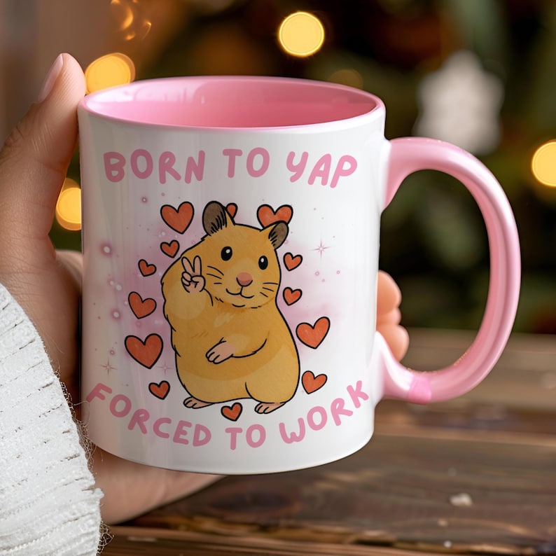 May include: White ceramic mug with a pink interior and handle. The mug features a cartoon hamster giving a peace sign, surrounded by hearts, and the text "BORN TO YAP FORCED TO WORK".