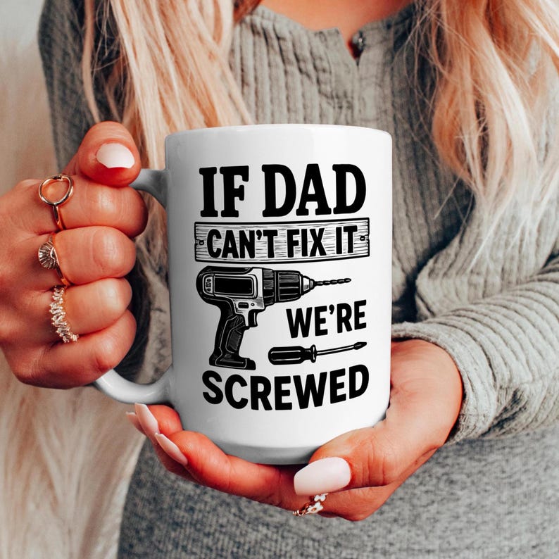 May include: White ceramic mug with the text "IF DAD CAN'T FIX IT WE'RE SCREWED" in black. The mug features a graphic of a drill and a screwdriver. The mug is held by a person.