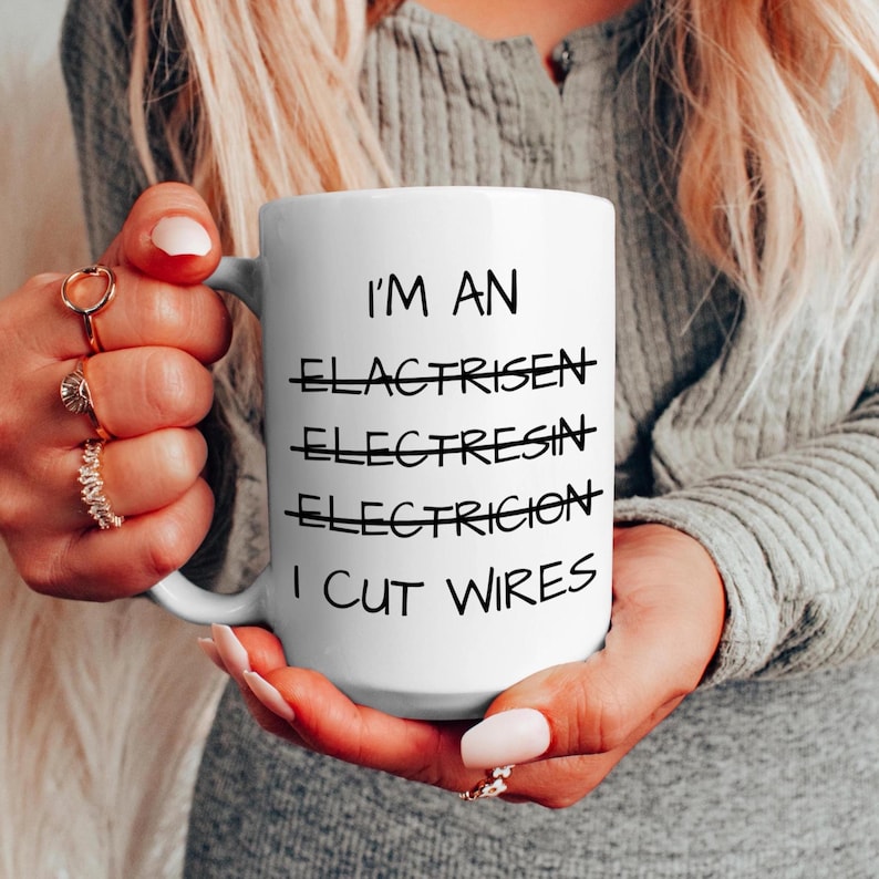 May include: A white ceramic mug featuring black text that reads I'M AN, followed by three crossed-out misspelled words, and finally the phrase I CUT WIRES. The mug has a smooth finish and a curved handle, designed for holding hot or cold beverages.