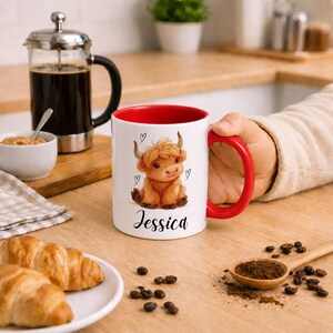 Pode incluir: Caneca de cer&acirc;mica branca com interior e al&ccedil;a vermelhos, com uma ilustra&ccedil;&atilde;o de vaca Highland e o nome "Jessica". A caneca &eacute; segurada por uma pessoa, com caf&eacute;, croissants e gr&atilde;os de caf&eacute; em uma superf&iacute;cie de madeira.